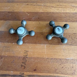 May include: Two vintage faucet handles with a four-petal design. One handle is labeled "COLD" and the other "HOT" in white text on a light blue background. The handles are made of a tarnished metal and rest on a wooden surface.