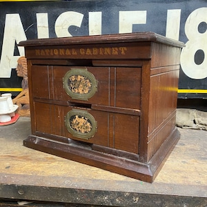 May include: A vintage wooden cabinet with two drawers, featuring brass-toned oval accents and the text "NATIONAL CABINET" on the top. The cabinet is a dark brown color and sits on a wooden surface.