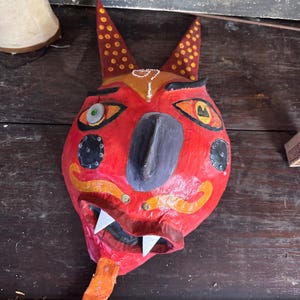 May include: A vibrant, handmade mask with a red base color. The mask features orange, yellow, black, and white accents, including pointed ears, stylized eyes, and sharp teeth. The mask has a long, orange tongue and is likely used for decorative or ceremonial purposes.