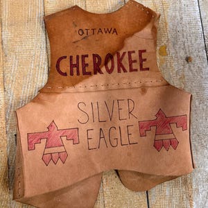 May include: A tan leather vest featuring the text OTTAWA and CHEROKEE in dark lettering at the top. Below, the text SILVER EAGLE is centered between two red-outlined bird motifs. The garment is constructed with visible stitching along the seams.