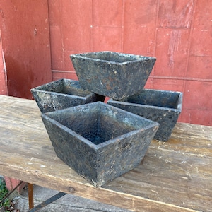 May include: Four square black planters made of rough textured material. The planters are stacked on top of each other on a wooden table.