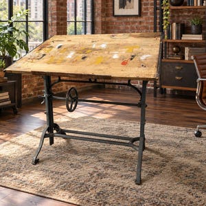 May include: A vintage drafting table with a weathered, light-colored wooden top featuring scattered paint splatters in yellow, white, and red. The table is supported by a dark, heavy-duty cast iron base with an adjustable hand crank mechanism.