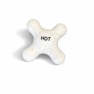 May include: A white, cross-shaped faucet handle with the word "HOT" printed in black. The handle has a textured surface and is likely made of ceramic or porcelain. The design is simple and functional.