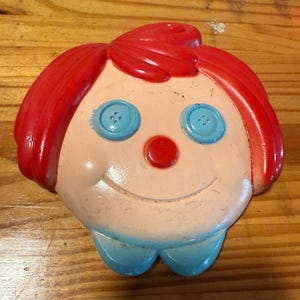 May include: A vintage, smiling face wall decoration. The face has red hair, blue button eyes, a red nose, and a blue bow tie. The face is painted on a light pink background.