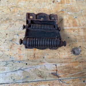 May include: A vintage, cast iron hinge with a spring mechanism. The hinge is dark brown with a weathered appearance, showing signs of age and use. It features a rectangular frame and two cylindrical handles. The spring is visible.