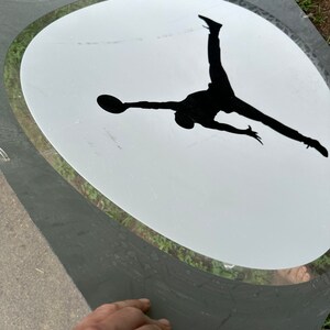 May include: A black silhouette of a person jumping in mid-air with a basketball in their hand, inside a white oval with a gray border.