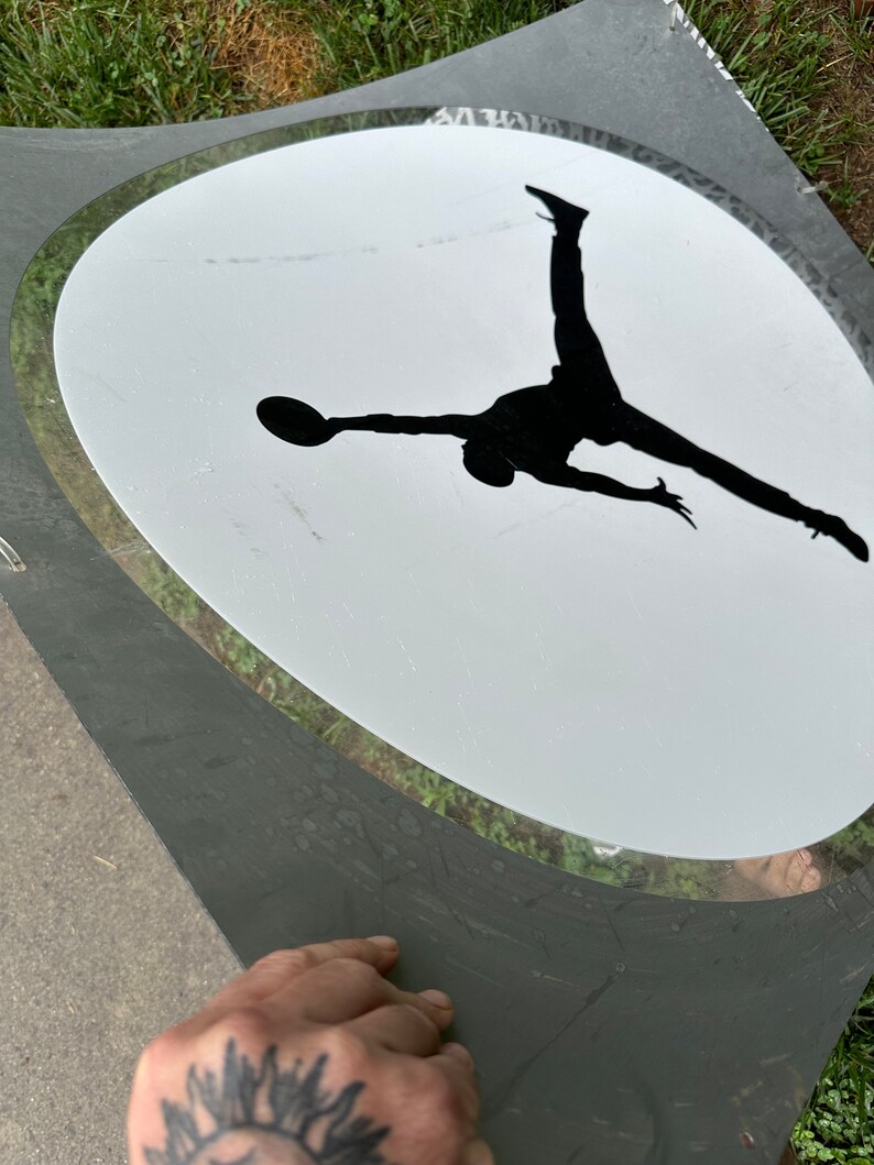 May include: A black silhouette of a person jumping in mid-air with a basketball in their hand, inside a white oval with a gray border.
