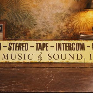 May include: A vintage-style sign with the text "AM-FM - STEREO - TAPE - INTERCOM - CHIMES by MUSIC & SOUND, Inc" in black letters on a yellow background. The sign is placed on a wooden surface, with a radio and other items in the background.