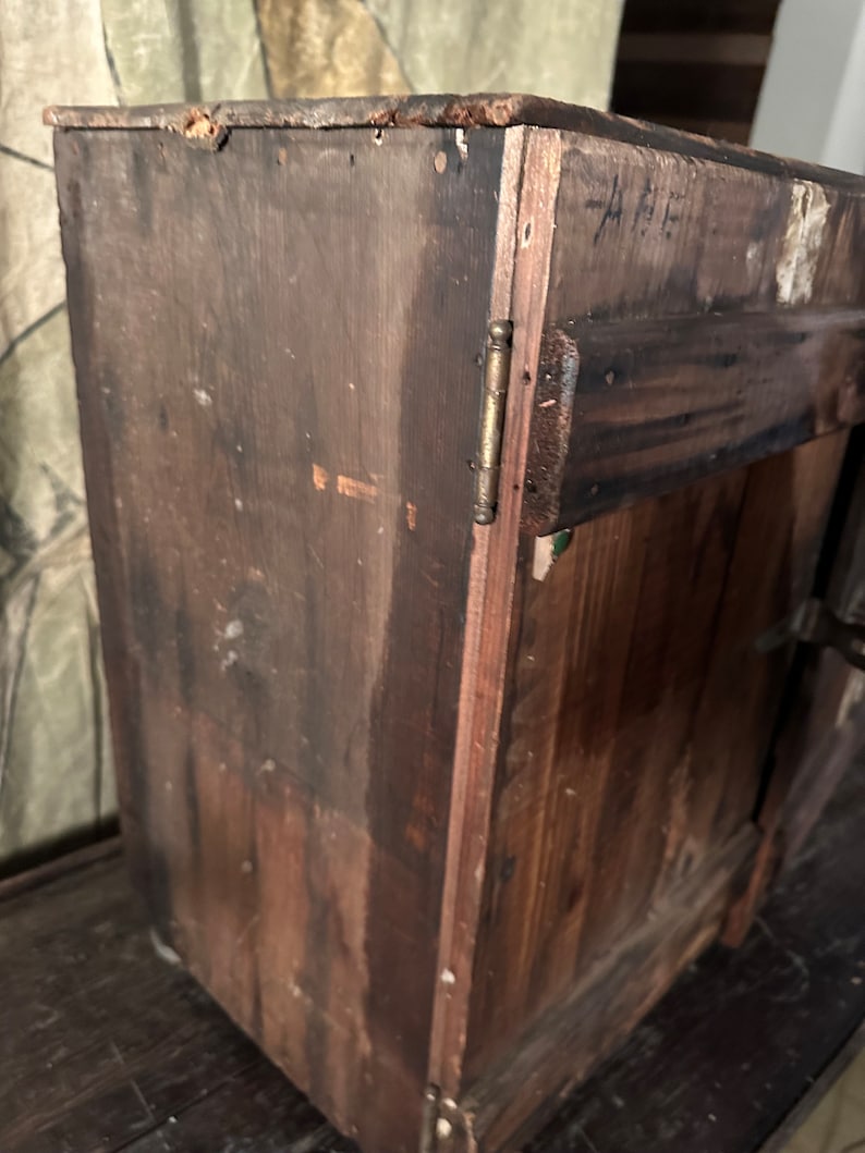 May include: A dark brown wooden cabinet with a single door and a brass hinge. The cabinet has a handwritten inscription on the side that reads "A.F."