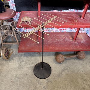 May include: A metal display stand featuring a black circular base and a vertical pole supporting a horizontal brass-toned rack. The rack consists of multiple extending arms with rounded tips, designed for hanging items in a tiered arrangement.
