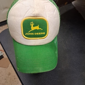 May include: A green and white trucker hat featuring a rectangular patch on the front with a yellow border. The patch displays a yellow leaping deer silhouette above the text JOHN DEERE in yellow. The hat has a green brim and mesh back panels.