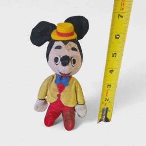 K&ouml;nnte beinhalten: Ein Vintage-Mickey-Mouse-Pl&uuml;schtier, etwa 15 cm gro&szlig;, mit gelber Jacke, roter Hose und gelbem Hut. Das Spielzeug hat schwarze Ohren und eine blaue Fliege. Ein gelbes Ma&szlig;band liegt neben dem Spielzeug.
