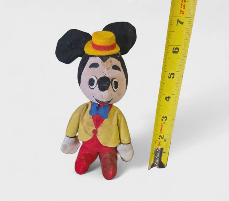 K&ouml;nnte beinhalten: Ein Vintage-Mickey-Mouse-Pl&uuml;schtier, etwa 15 cm gro&szlig;, mit gelber Jacke, roter Hose und gelbem Hut. Das Spielzeug hat schwarze Ohren und eine blaue Fliege. Ein gelbes Ma&szlig;band liegt neben dem Spielzeug.