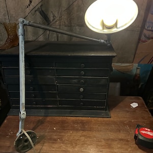 May include: A vintage industrial style lamp with a white metal arm and a round, white metal shade. The lamp is mounted on a black wooden drawer unit.