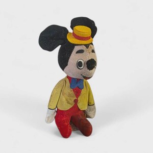 K&ouml;nnte beinhalten: Ein Vintage-Pl&uuml;schtier einer Mausfigur, die eine gelbe Jacke, eine rote Hose und eine blaue Fliege tr&auml;gt. Die Maus hat schwarze Ohren und einen gelben Zylinder. Das Spielzeug ist vor einem wei&szlig;en Hintergrund platziert.