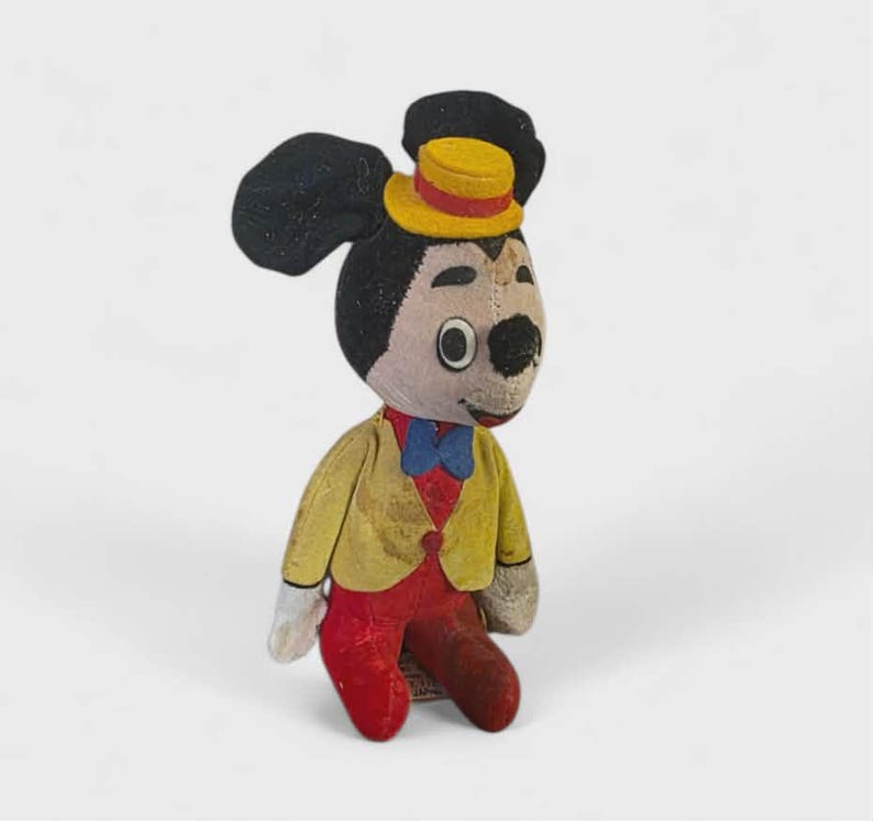 K&ouml;nnte beinhalten: Ein Vintage-Pl&uuml;schtier einer Mausfigur, die eine gelbe Jacke, eine rote Hose und eine blaue Fliege tr&auml;gt. Die Maus hat schwarze Ohren und einen gelben Zylinder. Das Spielzeug ist vor einem wei&szlig;en Hintergrund platziert.