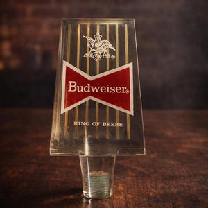 May include: Clear Budweiser tap handle with the Budweiser logo in red and white, and the words "KING OF BEERS". The handle has gold vertical stripes and a clear plastic base.