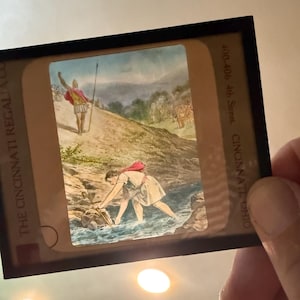 May include: A vintage slide featuring a historical scene with figures in classical attire. One figure stands on a hill with a spear, while another wades in a stream. The slide is held in a black frame.