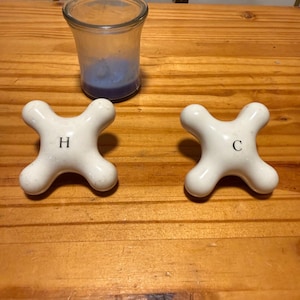 May include: Two white, cross-shaped faucet handles with smooth, rounded edges. One handle is marked with the black letter H, and the other is marked with the black letter C. A clear glass jar containing a blue substance is positioned behind them.