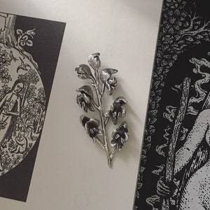 May include: A black and white illustration of a tree with two figures standing beneath it. The illustration is titled "The Root of Evil". A silver metal flower brooch is on a white surface.