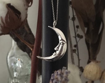 Puede incluir: Un collar con un colgante de media luna de plata con una cara esculpida en la luna. El collar cuelga de una cadena de plata y se muestra sobre un fondo de flores secas y una superficie oscura.