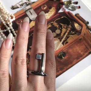 May include: A silver key-shaped ring with a square detail on the top. The ring is worn on a person's finger.