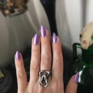 May include: A silver ring with a detailed design of a skull with a crown. The ring is worn on a hand with purple nail polish.