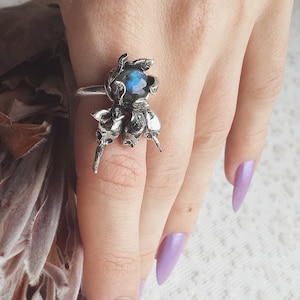 May include: A silver ring with a blue gemstone surrounded by delicate silver floral details. The ring is worn on a hand with light purple nail polish.