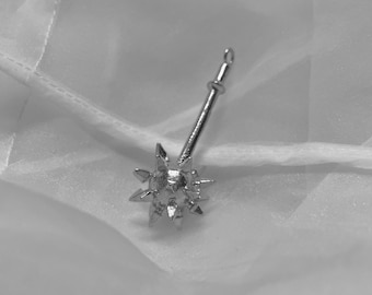 May include: A silver-colored earring with a starburst design. The earring has a long, thin post and a decorative head with multiple pointed projections. The earring is shown against a white, textured background.