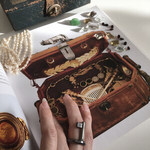 May include: A vintage wooden box with a brass latch, filled with various trinkets and tools. The box is open and a hand is wearing a silver ring with a geometric design.