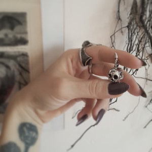 May include: A silver skull charm pendant with a detailed, textured surface. The pendant is hanging from a silver chain.