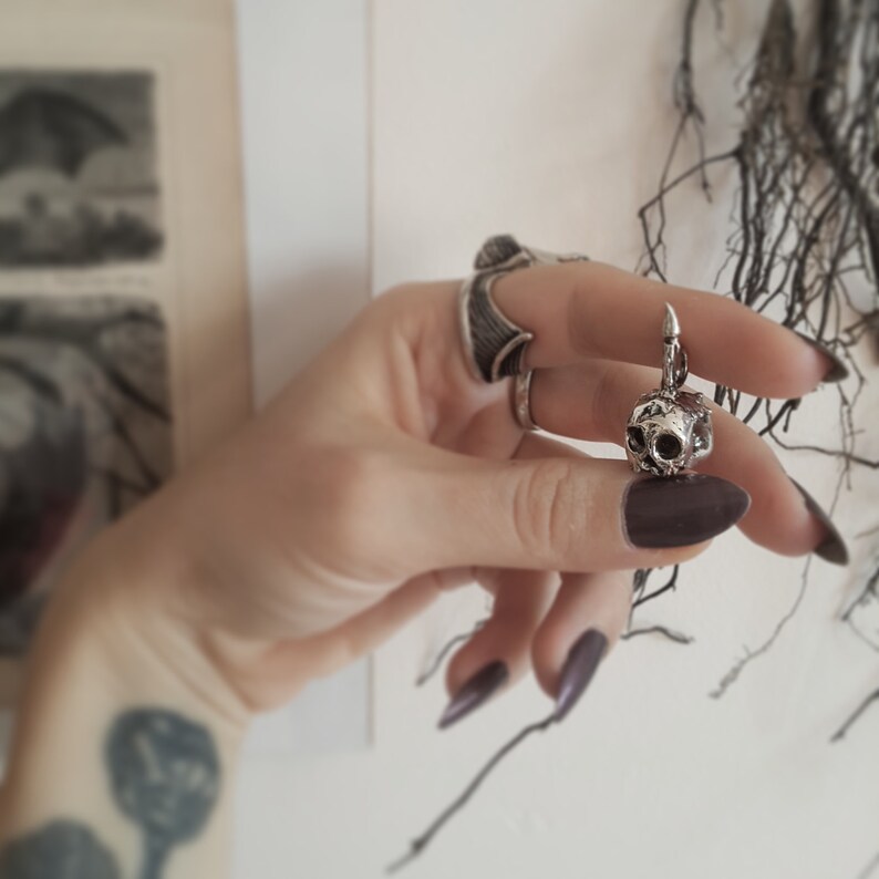 May include: A silver skull charm pendant with a detailed, textured surface. The pendant is hanging from a silver chain.