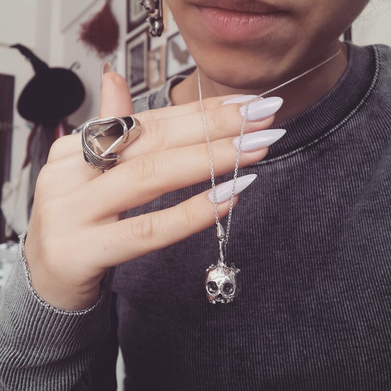 May include: A silver skull pendant necklace with a delicate chain. The necklace is worn by a person with white nail polish.