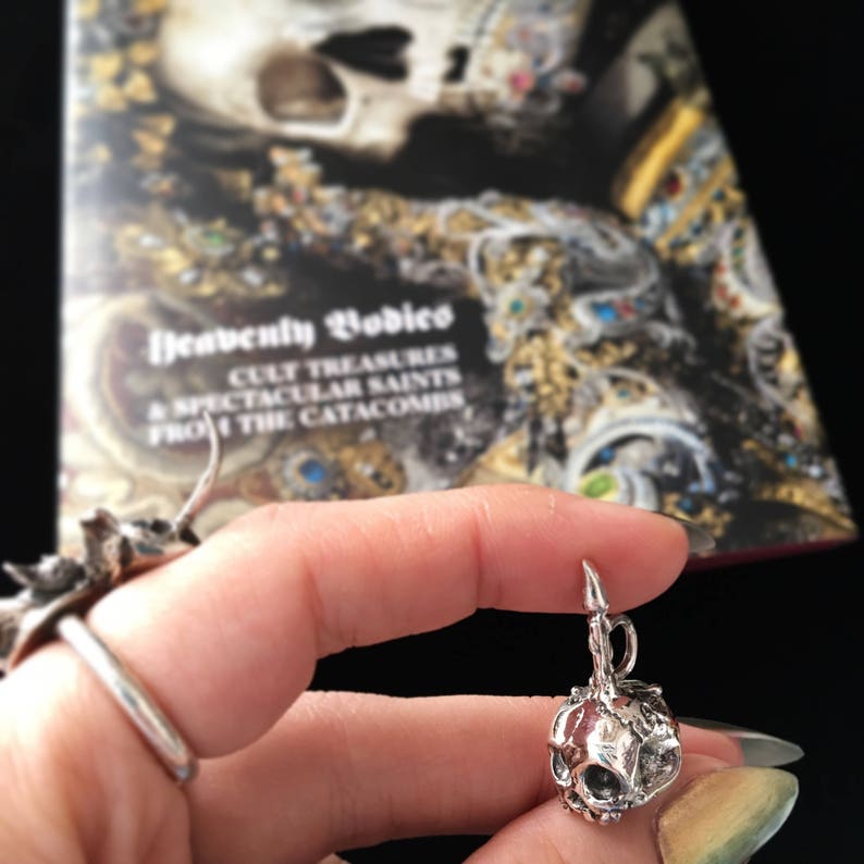 May include: Silver skull charm pendant with a detailed design. The skull is attached to a silver chain and has a small loop at the top for attaching to a necklace or bracelet.