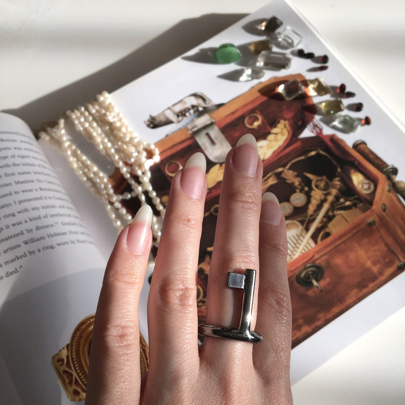May include: A silver key-shaped ring with a square detail on the top. The ring is worn on a person's finger.