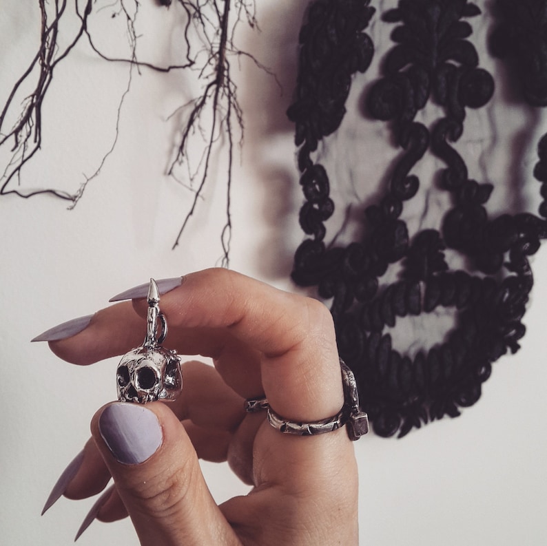 May include: A silver skull pendant with a pointed top, hanging from a chain. The pendant is being held by a hand wearing a silver ring.
