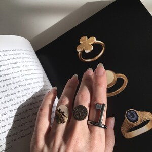 May include: A hand wearing a variety of rings, including a silver key-shaped ring, a gold ring with a dark stone, and a ring with a floral design. The rings are on a black background.
