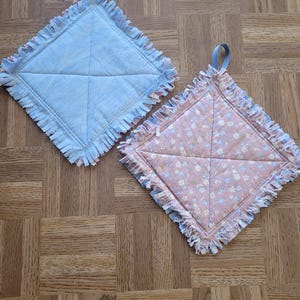 May include: Two square quilted pot holders. One is light blue with a blue fringe, the other is pink with a floral pattern and a blue fringe. Both have a blue hanging loop.