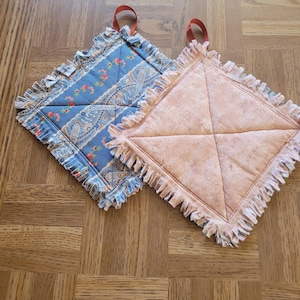 May include: Two square quilted pot holders with ruffled edges. One is blue with a floral and paisley pattern, the other is peach-colored. Both have a red loop for hanging.