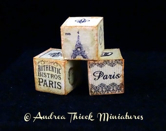 May include: Three wooden blocks stacked on top of each other. The top block has a black and white image of the Eiffel Tower and the text "Paris". The middle block has the text "Authentic Bistros Paris". The bottom block has a black and white image of a fleur-de-lis and the text "Paris".