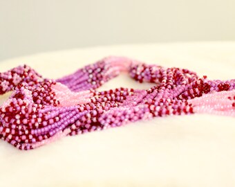 May include: A multi-strand beaded necklace in shades of pink, purple, and burgundy. The necklace is made of small, round beads, creating a textured and colorful accessory. The beads are arranged in a braided pattern.