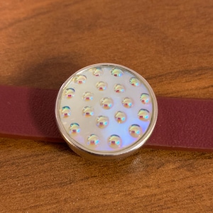May include: A round, silver-toned button with iridescent, bubble-like embellishments. The button is set on a burgundy leather strap, creating a bracelet-like appearance. The background is a warm, wooden surface.