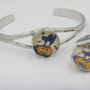 May include: A silver-toned cuff bracelet and two matching stud earrings. The bracelet and earrings feature a Halloween-themed design with a black cat on a jack-o'-lantern, with bats in the background. The jewelry is set against a white background.