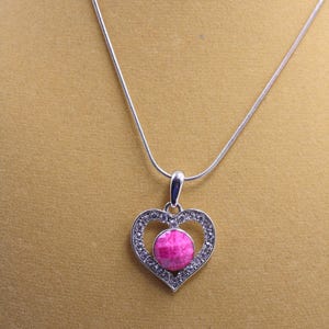 May include: A silver necklace with a heart-shaped pendant. The pendant has a pink gemstone and is encircled by small, clear crystals. The necklace has a fine chain.