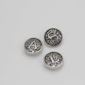 May include: Three silver-colored buttons with ornate designs and letters. The buttons are round with a raised border and a detailed pattern. The letters A, S, and L are featured on the buttons.