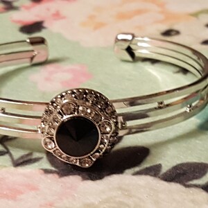 May include: Silver-toned cuff bracelet with a central black stone surrounded by clear crystals. The bracelet has a triple-band design with small decorative accents. The jewelry is displayed on a floral patterned surface.