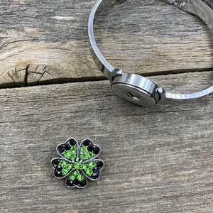 May include: A silver-toned bracelet with a snap closure and a decorative clover charm. The clover charm features green and black crystals, creating a sparkling accent. The bracelet is shown on a weathered wooden surface.