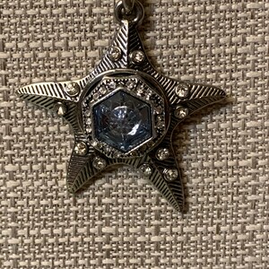 May include: Silver-toned star pendant necklace. The star has a textured design with small, clear crystals and a central, light blue, hexagonal stone. The necklace chain is a fine, silver-colored ball chain.