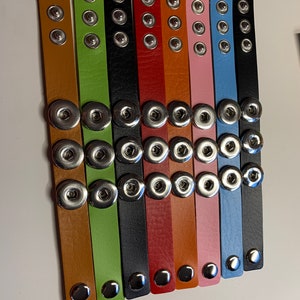 May include: A collection of colorful snap bracelets in various shades, including brown, green, black, red, orange, pink, blue, and gray. Each bracelet features three silver snap closures and is made of a smooth, leather-like material.