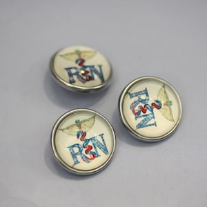 May include: Three round, silver-rimmed buttons with a white background. Each button features a blue and red caduceus symbol with the letters "RN" in blue, indicating Registered Nurse. The buttons are likely for medical professionals.